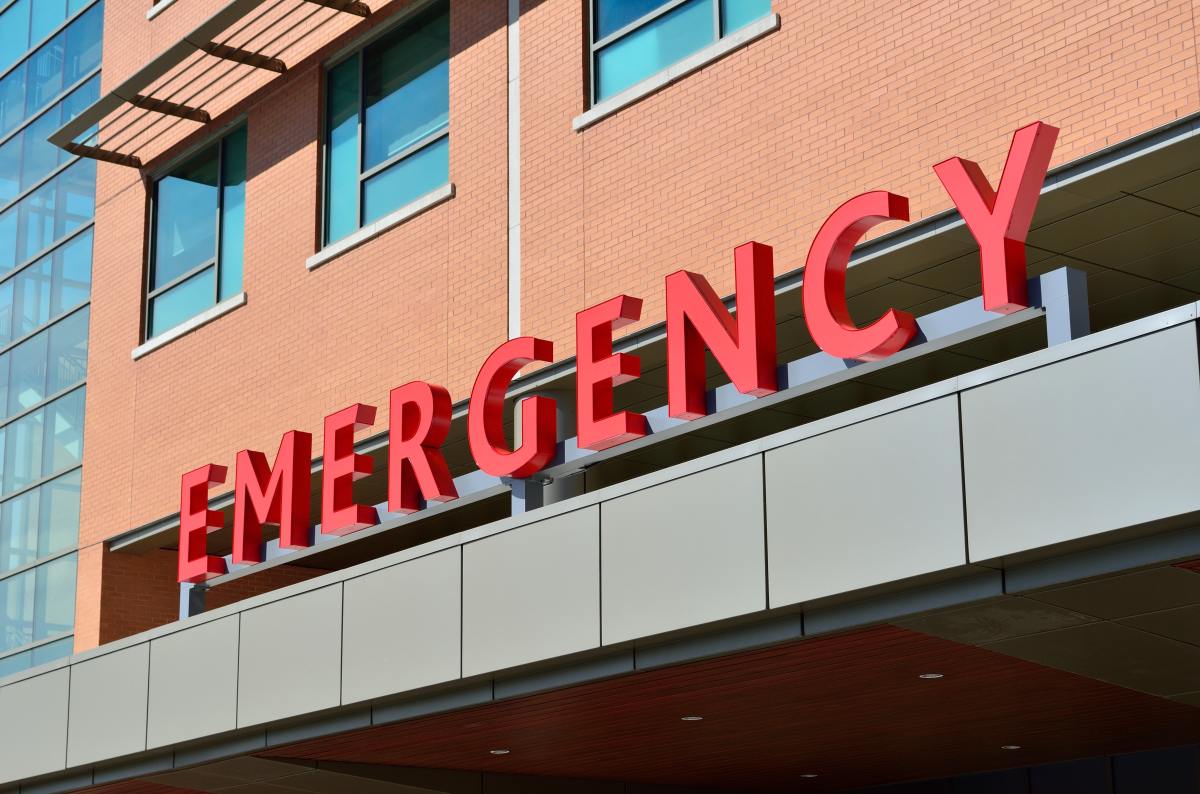 front of a brown hospital building and in red letters says emergency